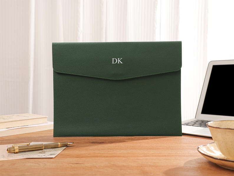 May include: A forest green document case with a flap, displaying the white initials "DK". It sits on a wooden desk, alongside a laptop, a pen, and a cup of tea.