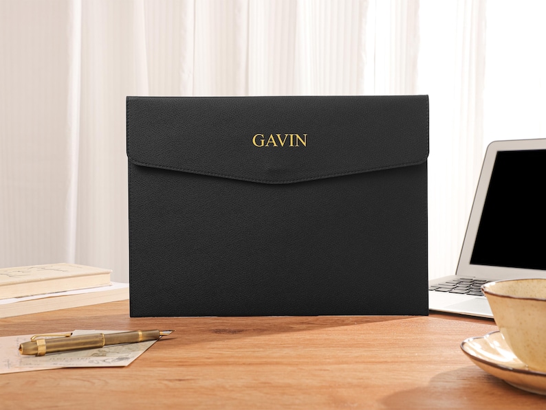 May include: Black laptop sleeve with the name "GAVIN" in gold lettering. The sleeve is on a wooden desk with a laptop, pen, and a cup of tea. The sleeve has a flap closure.