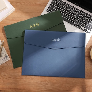 May include: Two personalised document holders. One is dark green with the initials "A.S.H" in gold lettering. The other is blue-grey with the name "Louis" in silver lettering. They are on a wooden surface.