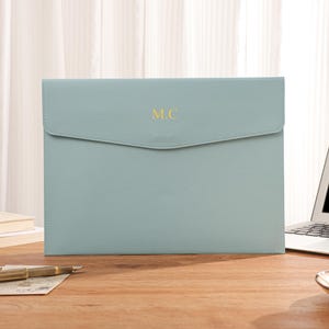 May include: A pale blue laptop sleeve with a flap closure. The initials "M.C" are embossed in gold on the front. A gold pen and a laptop are on a wooden desk, suggesting an office or workspace setting.