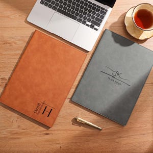 May include: Two personalised leather folios, one tan and one grey, are on a wooden surface beside a laptop and a teacup. The tan folio is inscribed with "Hazel" and the letter "H". The grey folio has the initials "JK" and the date "11.09.2025".