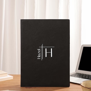 Könnte beinhalten: Ein schwarzes Notizbuch mit dem Namen "Hazel" und dem Buchstaben "H" in weißer, stilisierter Schrift. Das Notizbuch liegt auf einem Holzschreibtisch mit Laptop, Stift und Kaffeetasse. Im Hintergrund ein weißer Vorhang.