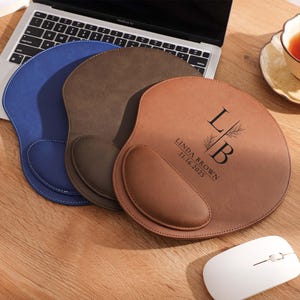 Puede incluir: Tres alfombrillas de ratón ergonómicas en azul, marrón y beige, con un portátil y una taza de té en el fondo. La alfombrilla beige tiene las iniciales "LB" y el nombre "LINDA BROWN" con la fecha "11.16.2025" impresa. También se ve un ratón blanco.
