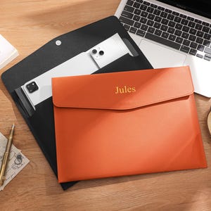 Puede incluir: Una funda para portátil de cuero naranja con el nombre "Jules" grabado en dorado. Una funda de cuero negro contiene una tableta blanca. Un portátil, un bolígrafo y una taza de té también están sobre la superficie de madera.