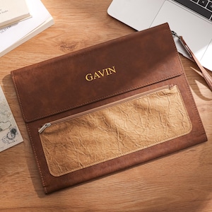 May include: A brown leather laptop sleeve with a zippered pocket and a wrist strap. The name "GAVIN" is embossed in gold lettering on the front. The sleeve is on a wooden surface, next to a laptop and books.