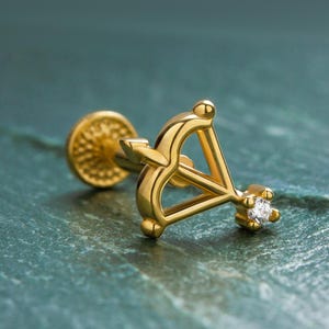 May include: A gold-coloured stud earring with a bow and arrow design, and a small, clear gemstone at the arrow's tip. The earring has a flat, textured back. The jewellery is set against a teal-coloured surface.