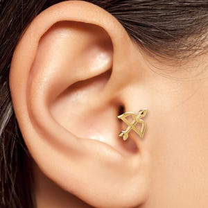 May include: A gold-coloured earring in the shape of a bow and arrow is shown on a person's ear. The earring is a small, delicate piece of jewellery with a detailed design. The earring is a stud style.