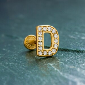 May include: A gold-coloured stud earring shaped like the letter "D", set with small, clear gemstones. The earring features a post and a textured backing. The background is a dark teal-green colour.