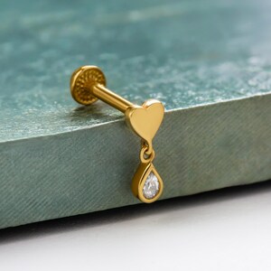 May include: A gold-colored lip piercing stud with a heart-shaped charm and a dangling teardrop-shaped gem. The stud has a flat, textured back and a straight post. The teardrop gem is clear and faceted.