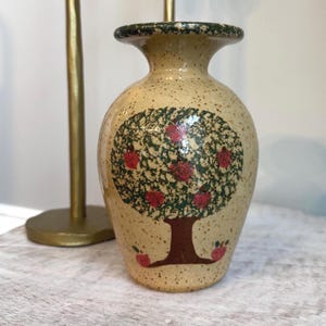 May include: A ceramic vase with a tree design featuring red apples. The vase has a speckled beige background, a dark green rim, and a brown tree trunk. Two gold candle holders are in the background.