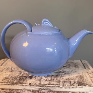 以下が含まれることがあります： 丸みを帯びたボディ、湾曲したハンドル、注ぎ口を備えた、淡い青色のセラミックティーポット。 蓋には装飾的なハンドルが付いています。 ティーポットは、使い古された木製の表面に置かれています。