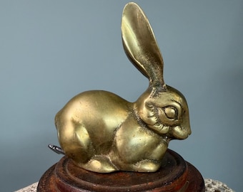 May include: A polished brass figurine of a rabbit in a seated position with one long ear pointed upward and the other folded back. The metallic gold-toned sculpture features smooth, rounded contours and sits atop a dark, circular wooden base.
