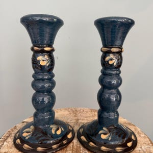 May include: A pair of dark blue ceramic candle holders with a glossy finish. Each holder features a flared top, a rounded central stem, and a wide circular base. The surface is decorated with hand-painted cream and dark brown leaf patterns.