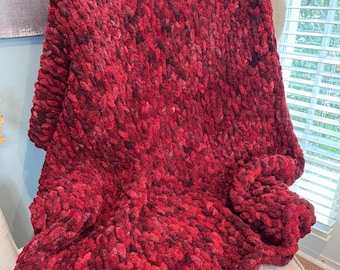May include: A chunky knit blanket in shades of red and burgundy is draped over a light-colored armchair. The blanket's texture is thick and plush, suggesting warmth and comfort. The cozy throw blanket is a home decor item.