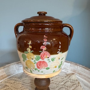 Puede incluir: Un tarro de cerámica marrón con tapa y asas, con un diseño floral en rosa, amarillo y verde. El tarro está sobre un soporte de madera, mostrando una estética vintage.