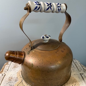 May include: A copper kettle with a white and blue floral patterned handle and lid knob. The kettle has a spout and a curved handle. The kettle is sitting on a white surface.