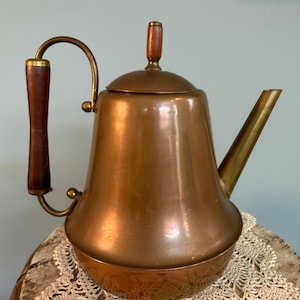 May include: A copper-colored teapot with a wooden handle and lid knob. The spout and handle are brass. The teapot sits on a decorative, off-white lace doily. The teapot has a bulbous body and a curved handle.