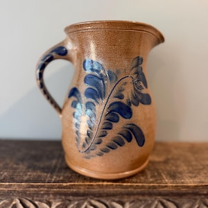 May include: A tan stoneware pitcher featuring a speckled texture and a hand-painted cobalt blue fern-like leaf pattern on the front and handle. The vessel has a wide rim, a curved spout, and a sturdy side handle, showcasing a rustic, earthy finish.