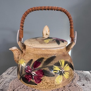 May include: Tan ceramic teapot with a woven handle and lid. The teapot is decorated with hand-painted flowers in yellow and burgundy. The handle is made of woven brown material. The teapot is sitting on a tan lace doily.