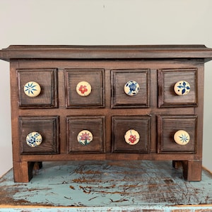 May include: A dark brown wooden chest with eight square drawers, each featuring a unique, hand-painted ceramic knob. The knobs display various floral and star-shaped patterns in blue, red, and green colors against a cream-colored background.