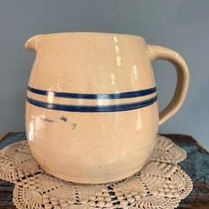 May include: A cream-colored ceramic pitcher featuring a rounded body, a curved handle, and a small pouring spout. Two horizontal dark blue stripes wrap around the upper portion of the pitcher, which has a smooth, glazed finish and a sturdy base.