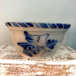 May include: A beige ceramic bowl featuring a hand-painted blue bird design on the exterior. The bowl has a speckled texture and a decorative blue rope-style pattern around the upper rim. The bird is depicted perched on a leafy branch.