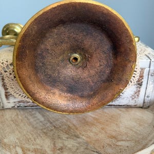 Puede incluir: Primer plano de la base de un candelabro de lat&oacute;n. La base es circular con un interior marr&oacute;n oscuro y un borde dorado. El centro tiene un peque&ntilde;o agujero para un tornillo. El candelabro est&aacute; sobre una superficie de madera.
