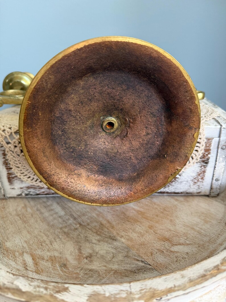 Puede incluir: Primer plano de la base de un candelabro de lat&oacute;n. La base es circular con un interior marr&oacute;n oscuro y un borde dorado. El centro tiene un peque&ntilde;o agujero para un tornillo. El candelabro est&aacute; sobre una superficie de madera.