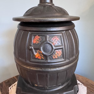 Könnte beinhalten: Ein dunkelbrauner Behälter aus Gusseisen im antiken Stil mit Deckel. Der Behälter hat eine Fassform mit dekorativen Paneelen und orangefarbenen und roten Akzenten. Er steht auf einem rechteckigen Sockel und dient wahrscheinlich als dekoratives Aufbewahrungsstück.