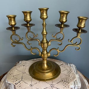 Puede incluir: Un candelabro de lat&oacute;n con cinco candeleros. El candelabro tiene una base redonda y brazos curvos. Los candeleros tienen forma de copa. El candelabro es de lat&oacute;n y tiene un acabado brillante. Es un objeto decorativo.