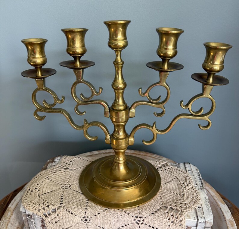 Puede incluir: Un candelabro de lat&oacute;n con cinco candeleros. El candelabro tiene una base redonda y brazos curvos. Los candeleros tienen forma de copa. El candelabro es de lat&oacute;n y tiene un acabado brillante. Es un objeto decorativo.