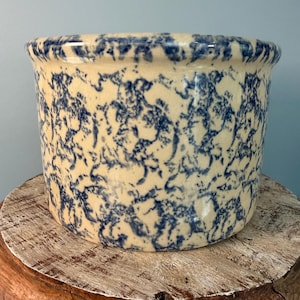 May include: A round, ceramic stoneware crock with a cream-colored base and a blue, spattered design. The rim is slightly flared. The crock is sitting on a wooden pedestal with a distressed finish. The background is a solid light blue.