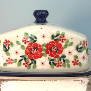 May include: A ceramic butter dish with a lid, featuring a floral design with red and white flowers, green leaves, and a navy blue knob. The dish sits on a matching tray with a navy blue trim, perfect for kitchen decor.