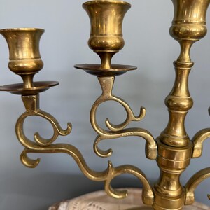 Puede incluir: Un candelabro de lat&oacute;n con tres candeleros. El candelabro tiene un dise&ntilde;o decorativo y curvo con una base redonda. Los candeleros tienen forma de peque&ntilde;as copas. El lat&oacute;n tiene un color dorado c&aacute;lido.