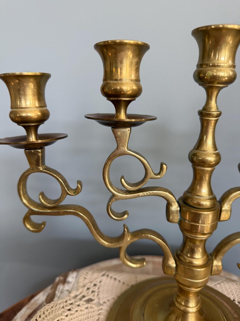 Puede incluir: Un candelabro de lat&oacute;n con tres candeleros. El candelabro tiene un dise&ntilde;o decorativo y curvo con una base redonda. Los candeleros tienen forma de peque&ntilde;as copas. El lat&oacute;n tiene un color dorado c&aacute;lido.