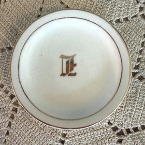 May include: A small, round, white dish with a gold rim and a gold letter "E" in the center. The dish has a slightly cracked surface and is set against a textured, beige background.