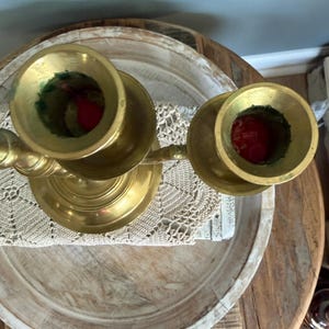 Puede incluir: Un candelabro de lat&oacute;n con dos candeleros. Los candeleros son circulares y tienen una base ancha. El candelabro est&aacute; colocado sobre una bandeja de madera con un tapete de encaje blanco. Las velas son rojas.