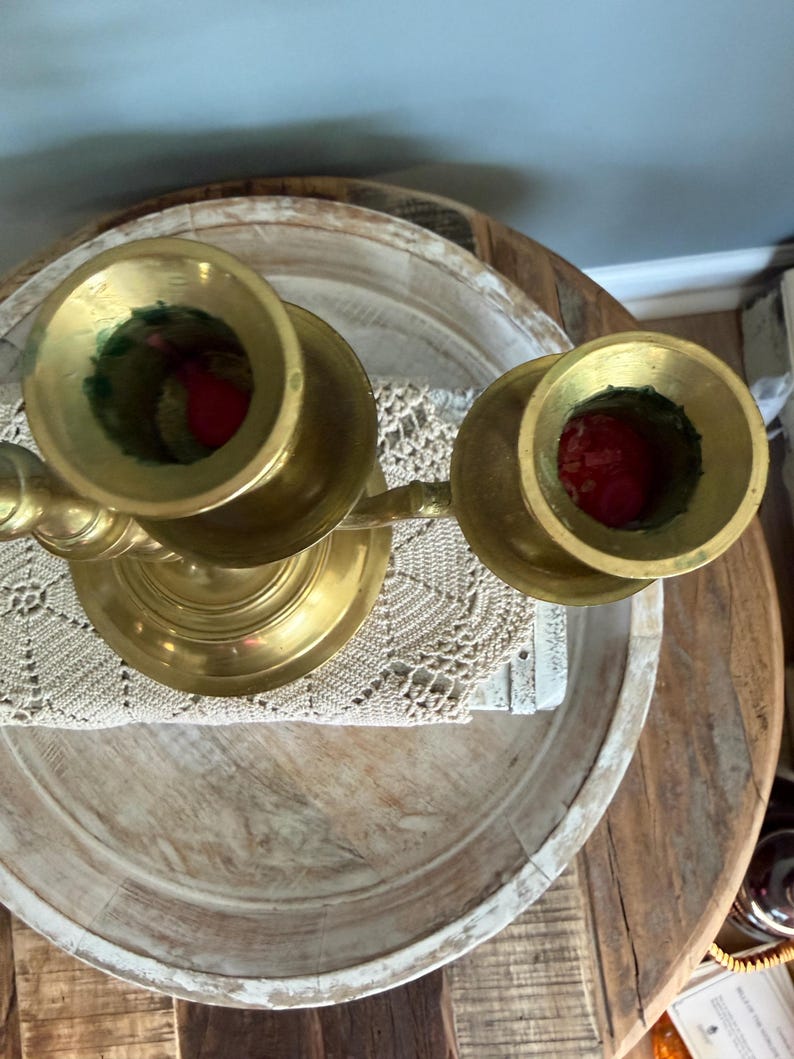 Puede incluir: Un candelabro de lat&oacute;n con dos candeleros. Los candeleros son circulares y tienen una base ancha. El candelabro est&aacute; colocado sobre una bandeja de madera con un tapete de encaje blanco. Las velas son rojas.