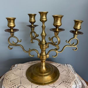 Puede incluir: Un candelabro de lat&oacute;n vintage con cinco candeleros. El candelabro tiene una base redonda y brazos curvos ornamentados. Los candeleros est&aacute;n dise&ntilde;ados para sostener velas de tama&ntilde;o est&aacute;ndar. El lat&oacute;n tiene un tono dorado c&aacute;lido.