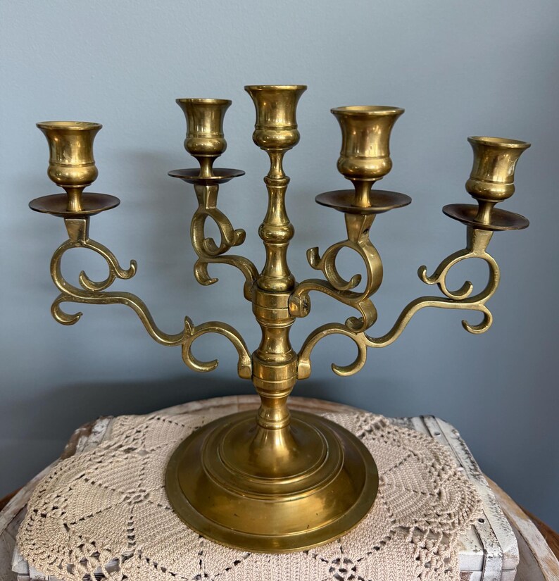 Puede incluir: Un candelabro de lat&oacute;n vintage con cinco candeleros. El candelabro tiene una base redonda y brazos curvos ornamentados. Los candeleros est&aacute;n dise&ntilde;ados para sostener velas de tama&ntilde;o est&aacute;ndar. El lat&oacute;n tiene un tono dorado c&aacute;lido.