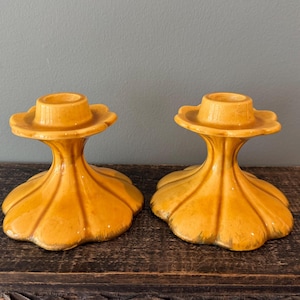 May include: A pair of matching mustard yellow ceramic candle holders featuring a wide, scalloped base that tapers upward to a circular top. The glazed finish has a smooth, reflective surface with subtle variations in the yellow color tone.