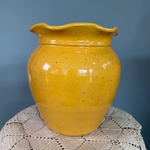 May include: A yellow ceramic vase with a ruffled rim. The vase has a rounded body and a speckled texture. The background is a solid blue color. The vase is sitting on a cream-colored, crocheted surface.