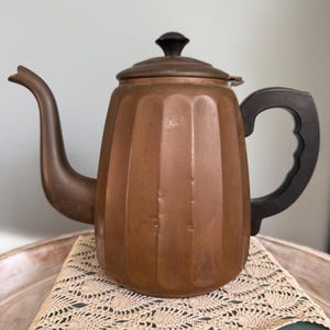 May include: A vintage copper teapot with a black handle and lid knob. The teapot has a ribbed design and a curved spout. It sits on a cream-colored lace doily.