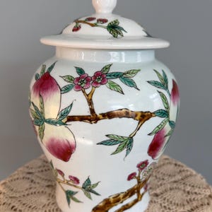 Pode incluir: Um pote de gengibre de cer&acirc;mica branca com tampa correspondente e um remate pontiagudo. O pote &eacute; decorado com ilustra&ccedil;&otilde;es pintadas &agrave; m&atilde;o de ramos de &aacute;rvore castanhos, folhas verdes, flores cor-de-rosa e p&ecirc;ssegos vermelhos e amarelos.