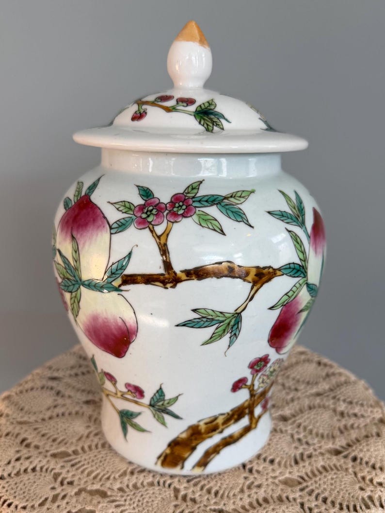 Pode incluir: Um pote de gengibre de cer&acirc;mica branca com tampa correspondente e um remate pontiagudo. O pote &eacute; decorado com ilustra&ccedil;&otilde;es pintadas &agrave; m&atilde;o de ramos de &aacute;rvore castanhos, folhas verdes, flores cor-de-rosa e p&ecirc;ssegos vermelhos e amarelos.