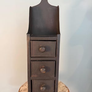 May include: A dark brown, vertical wall-mounted cabinet with three drawers and a decorative top. The cabinet has a curved back and a small shelf above the drawers. Each drawer has a round wooden knob. The cabinet is made of metal.