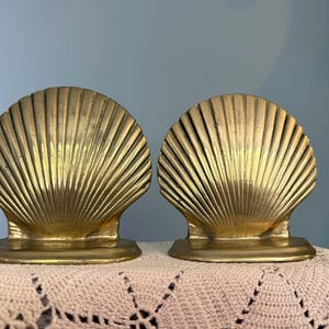 May include: A pair of gold-colored metal bookends, each shaped like a large, fan-like scallop shell with prominent vertical ridges radiating from the base to the rounded top edge, featuring a smooth, flat horizontal base for stability.