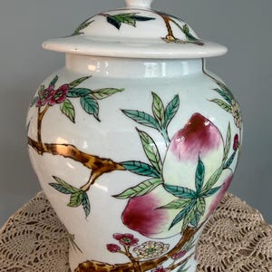 Pode incluir: Um pote de gengibre de cer&acirc;mica branca com tampa correspondente, apresentando ilustra&ccedil;&otilde;es pintadas &agrave; m&atilde;o de galhos de &aacute;rvore castanhos, folhas verdes, pequenas flores cor-de-rosa e grandes p&ecirc;ssegos em tons de rosa e creme. Acabamento liso e brilhante.