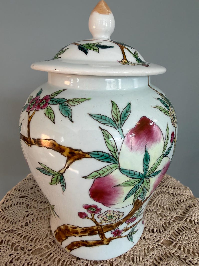 Pode incluir: Um pote de gengibre de cer&acirc;mica branca com tampa correspondente, apresentando ilustra&ccedil;&otilde;es pintadas &agrave; m&atilde;o de galhos de &aacute;rvore castanhos, folhas verdes, pequenas flores cor-de-rosa e grandes p&ecirc;ssegos em tons de rosa e creme. Acabamento liso e brilhante.