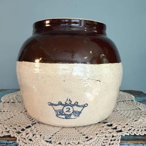 May include: A stoneware crock featuring a glossy dark brown glazed upper section and a cream-colored lower body. A blue stamped graphic of a crown with the number 2 inside is centered on the front of the vessel, which has a wide, rounded shape.